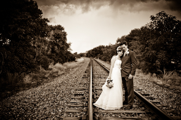 sepia image - a beautiful photo of the bride and groom embracing as they stand in the middle of a train track in the countryside - bride is wearing an a-line dress and birdcage veil - photo by New Mexico based wedding photographers Twin Lens